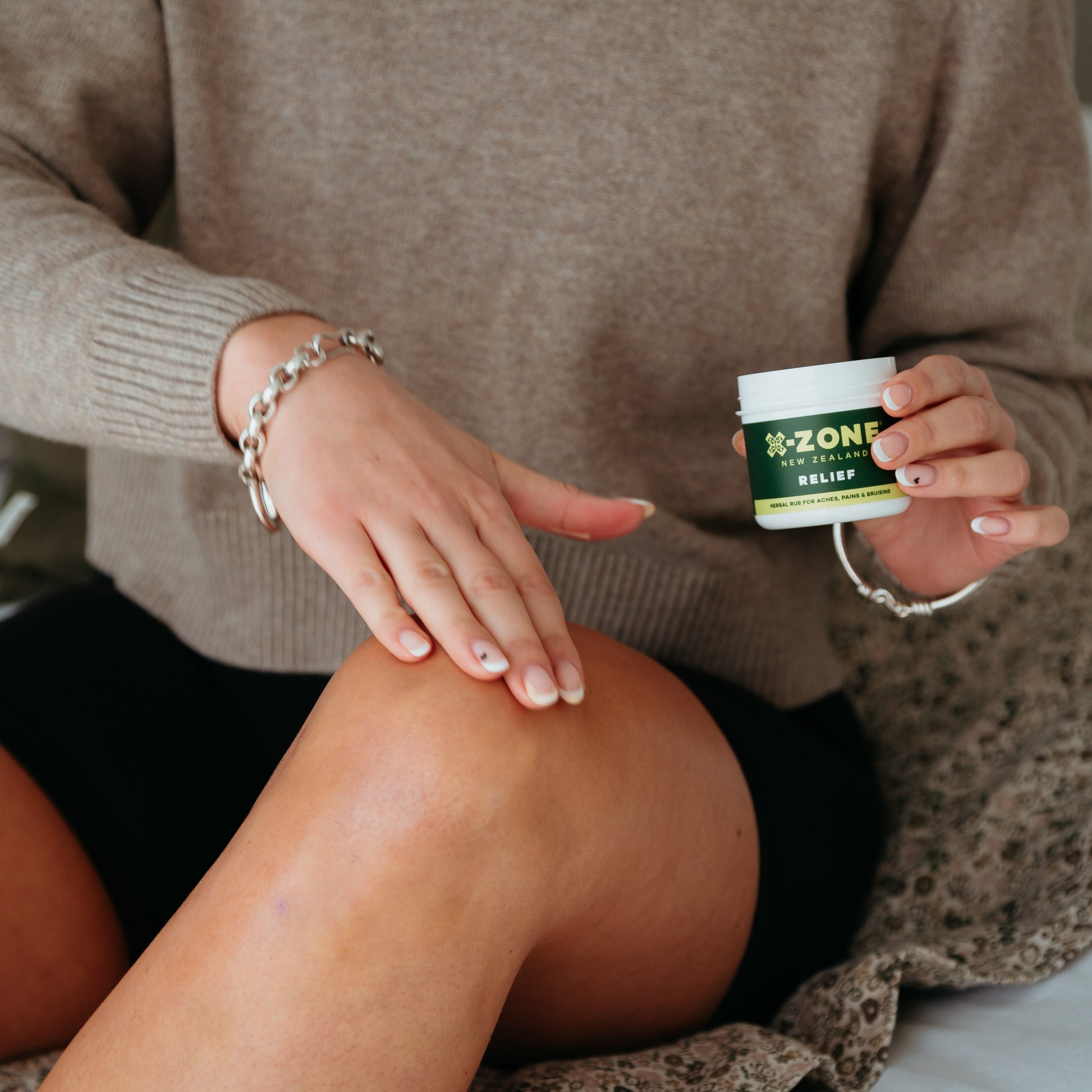 Person applying a cream to their knee with a container labeled X-ZONE RELIEF on a neutral background.