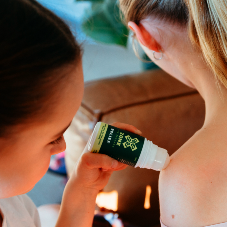 Person applying a roll on bottle labeled X-Zone relief to another person's shoulder.